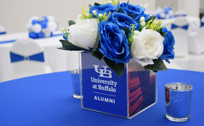 Table with a place setting of blue and white flowers in a cube that reads "University at Buffalo Alumni." Blue candles are on either side and there are decorated tables and chairs in the background.
