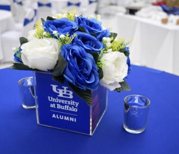 Place setting on a table of blue and white flowers in a cube that says "University at Buffalo alumni," with blue candles on either side