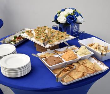 Table of appetizers including phyllo cup bites, hummus and pita chips, and caprese skewers. A place setting of white and blue flowers in a cube that reads "University at Buffalo alumni"